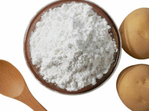 Top view of white powder in a bowl displayed with two potatoes and a wooden spoon.