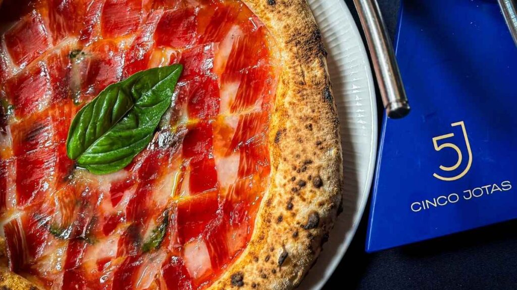 High-angle shot of a Margherita Royale pizza topped with thin, glossy slices of Cinco Jotas Ibérico ham, fresh basil, and buffalo mozzarella on a rustic wooden board.