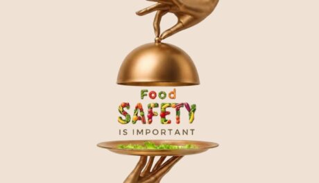 A golden hand lifts a cloche to reveal the words "Food Safety" spelled out in colorful fruits and vegetables, while another golden hand holds a platter of fresh greens below.