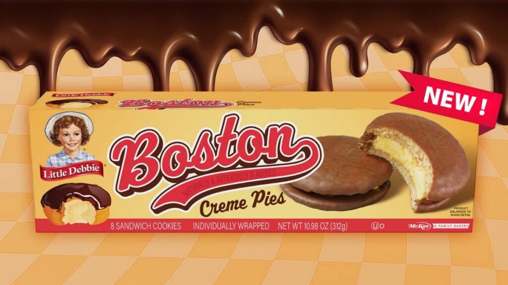 A box of Little Debbie Boston Creme Pie snacks, showing the product with a bite taken out to reveal the vanilla cookie and creme layers inside the chocolate coating.