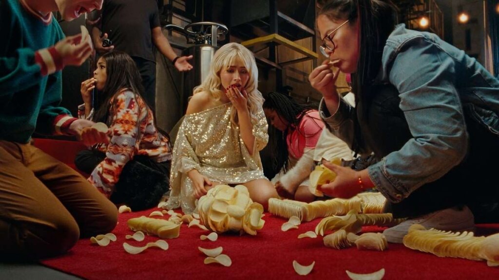A promotional shot for the 2026 Pringles Super Bowl ad featuring singer Sabrina Carpenter. She is holding a Pringle crisp next to "Pringleleo," an animatronic soulmate character constructed entirely out of stacked potato chips, set against a warm studio background.
