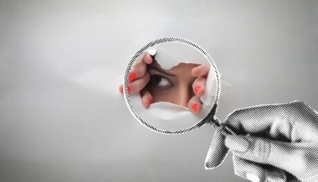 Eye peering through a tear in a sheet of paper while a magnifying glass examines it.