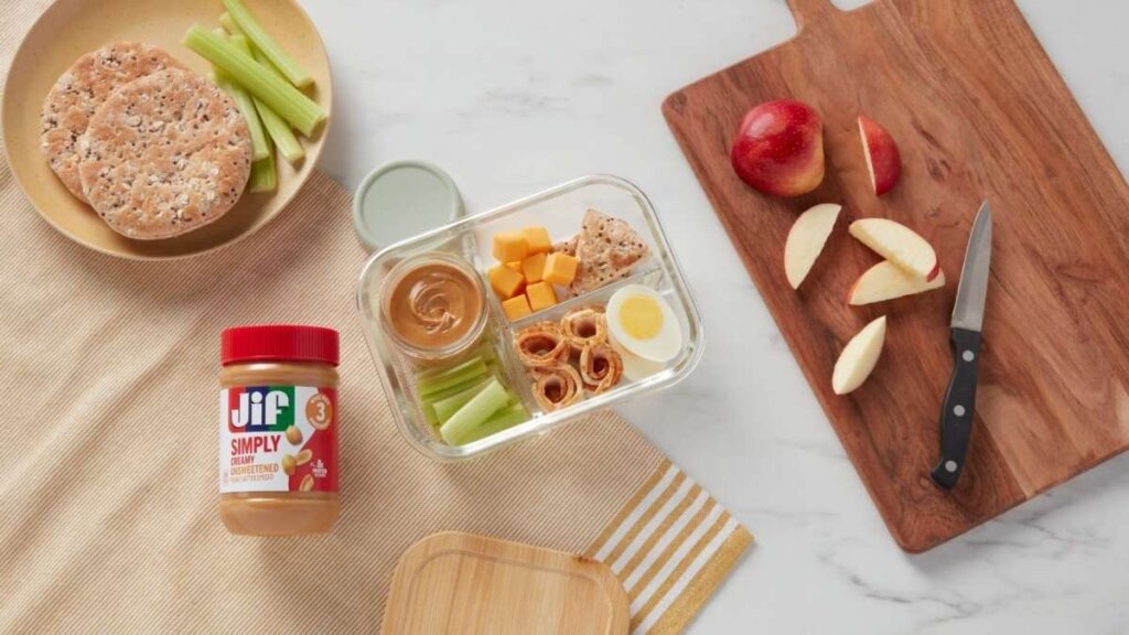 A jar of Jif Simply Unsweetened Creamy peanut butter next to a bowl of fresh roasted peanuts.